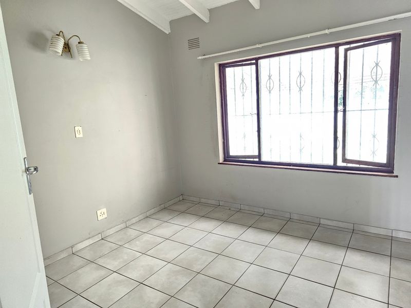 A vacant bedroom featuring tiled flooring, exposed beam ceiling, and a window fitted with security bars.