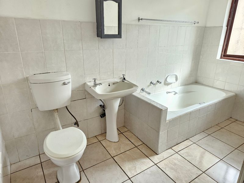 A dated bathroom featuring a pedestal basin, built-in bathtub, and toilet with neutral wall and floor tiling, offering potential for modernization.