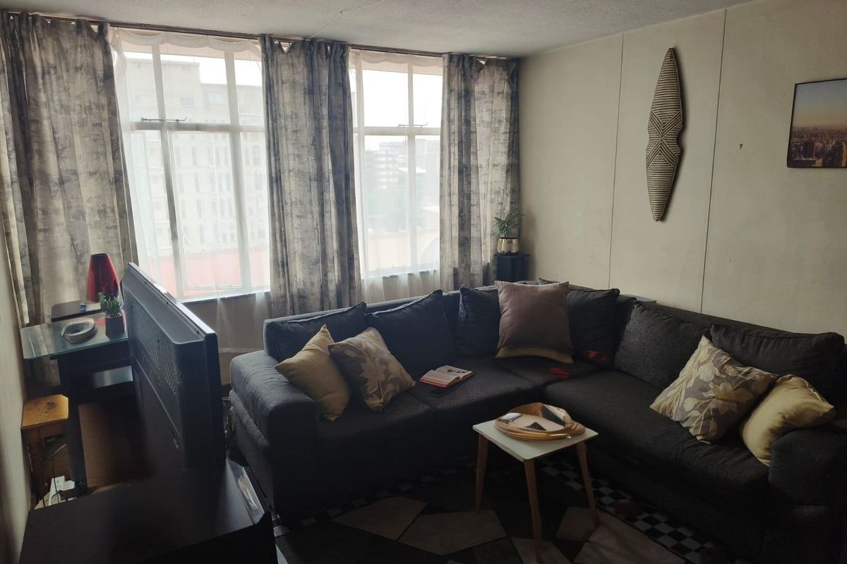 A living room featuring large windows that provide natural light, with wall and ceiling finishes showing signs of wear and age.