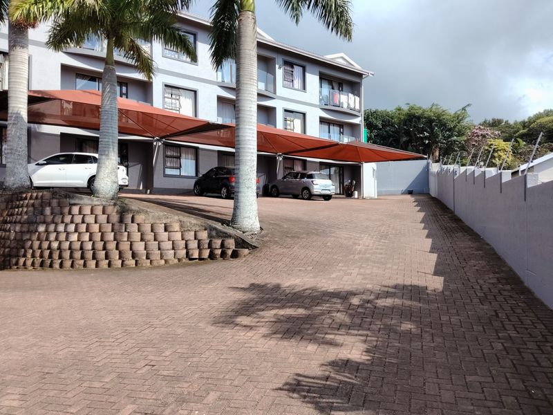 Exterior view of the apartment complex featuring a paved driveway, covered parking bays with shade cloth, and a secure perimeter wall.