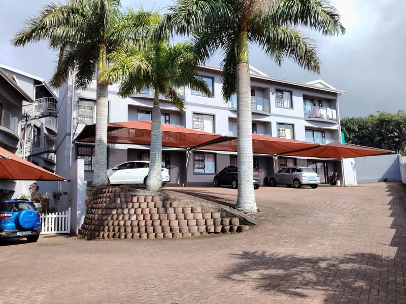 Front exterior view of a multi-story apartment building featuring a paved driveway, covered parking bays, and balconies with glass balustrades.