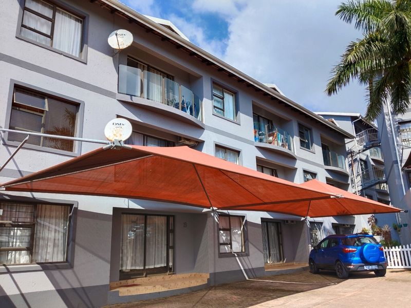 Exterior view of a multi-story apartment complex featuring balconies, sliding glass doors, and a large shade sail covering the paved parking area.