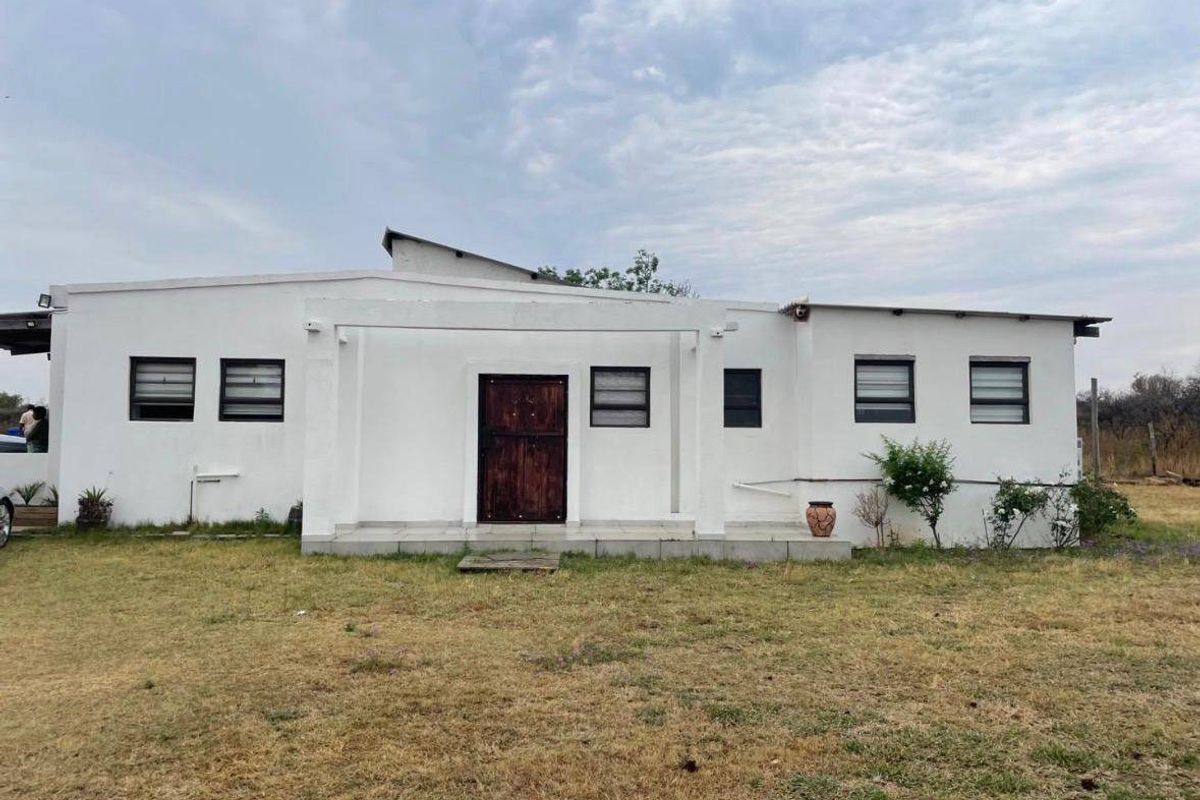 Rear view of a white-painted farm dwelling featuring a tiled patio area, timber entrance door, and multiple aluminium-framed windows.