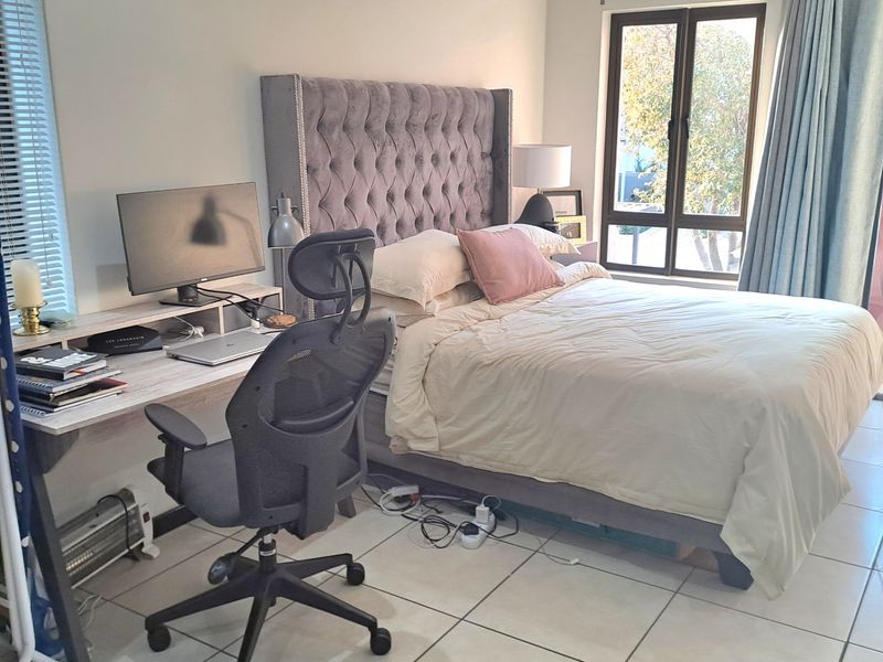 A well-lit bedroom featuring tiled flooring, neutral walls, and large windows with both blinds and curtains, providing a comfortable and functional living space.