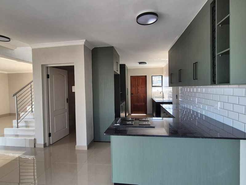 Modern open-plan kitchen featuring sage green cabinetry, granite countertops, subway tile backsplash, and a gas hob, flowing into an adjacent staircase area.