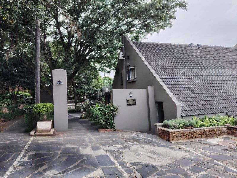 Front exterior view of the property featuring a steep tiled roof, stone retaining wall, and slate paved driveway area with mature trees.