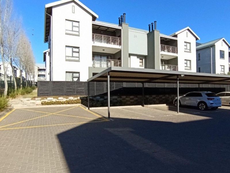 Exterior view of a multi-story apartment complex featuring covered parking bays, paved driveway, and private balconies for each unit.