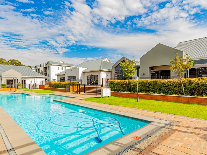 A well-maintained communal swimming pool area featuring paved decking, landscaped garden beds, and brick retaining walls within a modern apartment complex.