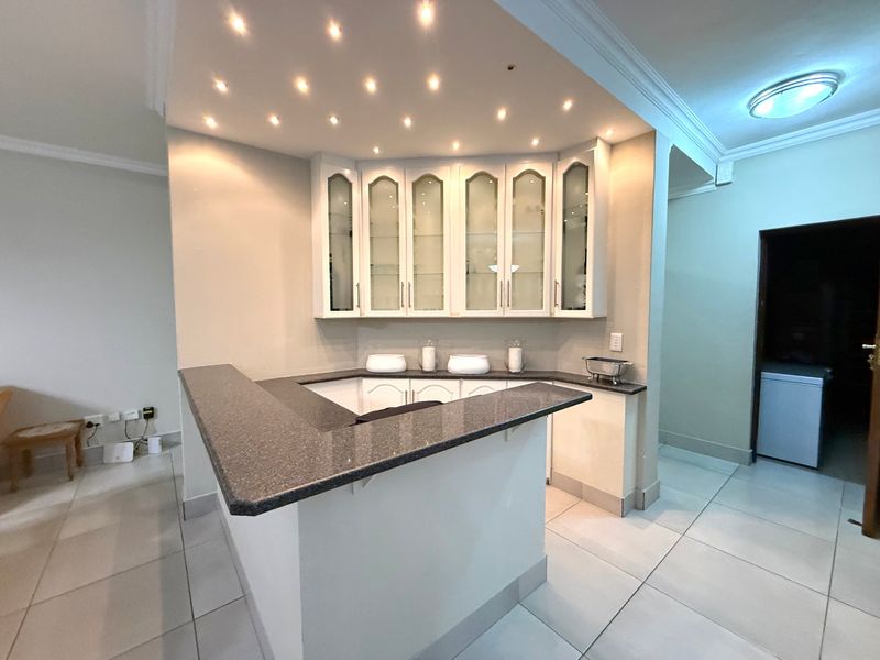 A dedicated built-in bar area featuring granite countertops, glass-fronted display cabinetry, and recessed downlighting, set within a tiled open-plan living space.