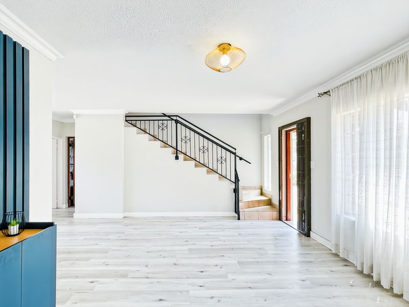 Spacious living area featuring light-toned laminate flooring, a central staircase with a decorative wrought iron balustrade, and neutral wall finishes.