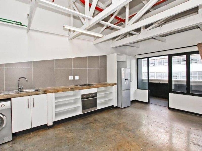 Open-plan kitchen and living area featuring industrial-style exposed roof trusses, concrete flooring, and direct access to a balcony through sliding glass doors.