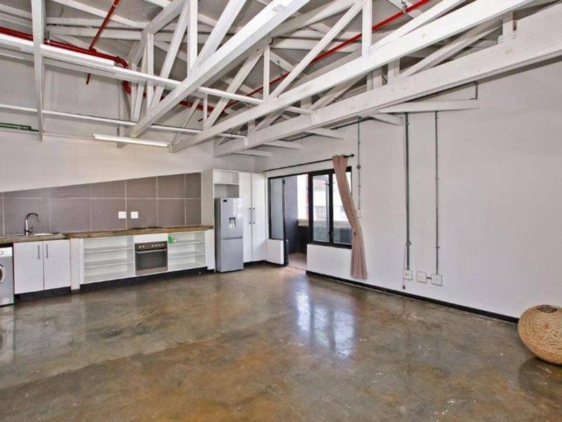 Spacious open-plan apartment featuring industrial-style exposed roof trusses, polished concrete floors, and a functional kitchen area with built-in cabinetry.