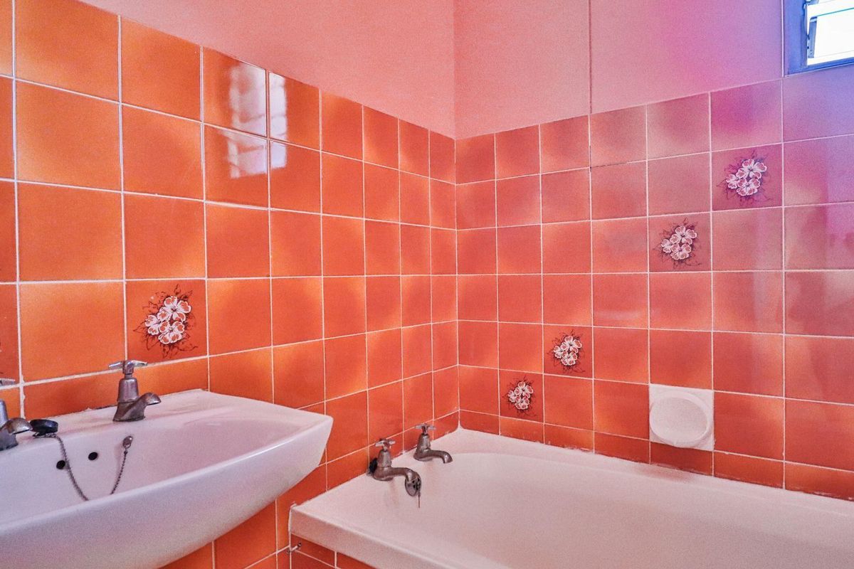 A dated bathroom featuring orange floral-patterned wall tiles, a white pedestal basin, and a built-in bathtub with chrome fixtures.