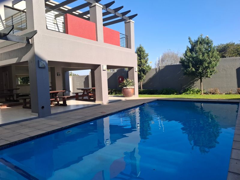 A well-maintained outdoor pool area featuring a clear swimming pool, tiled surround, and a covered patio with a pergola structure.