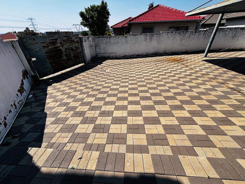 A paved outdoor area featuring a perimeter wall, metal gate, and carport structure, showing signs of wear and maintenance requirements.