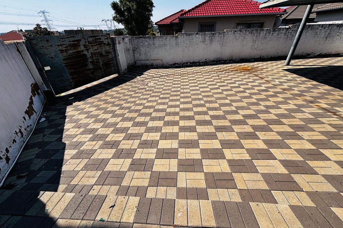 A paved outdoor area featuring a perimeter wall, metal gate, and carport structure, showing signs of wear and maintenance requirements.