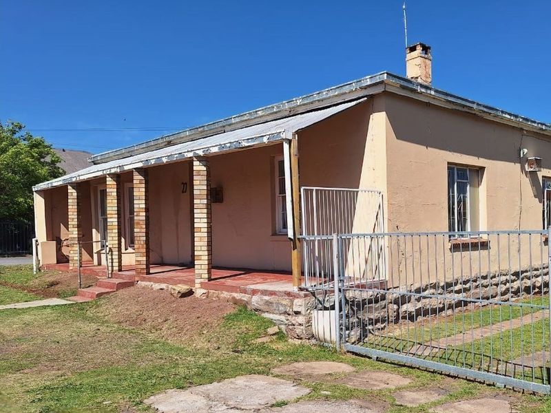 Front elevation of a single-story house featuring a covered veranda, stone foundation, and metal roofing, showing signs of age and exterior wear.