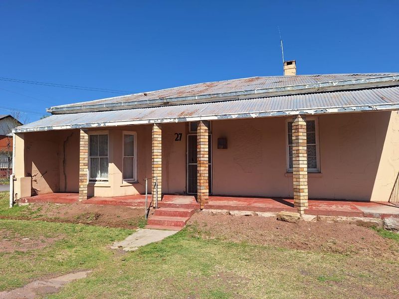Front elevation of a single-story house featuring a corrugated iron roof, brick pillars, and a covered veranda, showing signs of significant exterior wear.