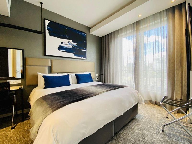 A well-maintained bedroom featuring a modern upholstered headboard, recessed ceiling lighting, and large sliding glass doors with floor-to-ceiling curtains providing natural light.