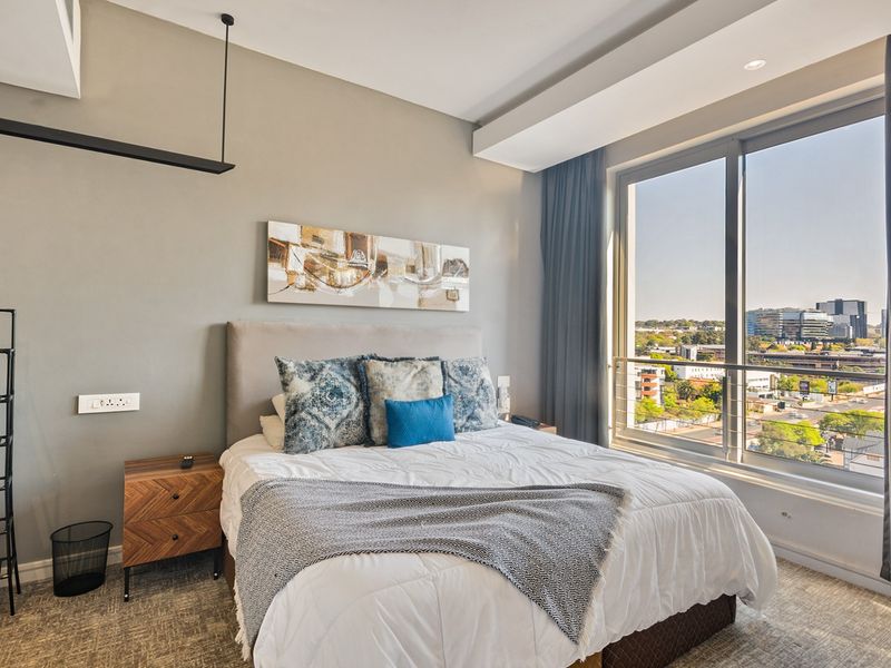 A modern bedroom featuring a large floor-to-ceiling window with expansive city views, neutral wall tones, and recessed ceiling lighting.