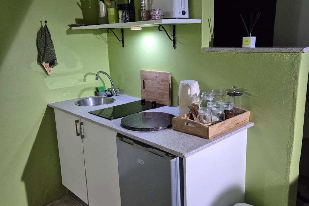 Compact kitchenette featuring white cabinetry, a stone-look countertop, integrated electric hob, stainless steel sink, and a convenient wall-mounted storage shelf.
