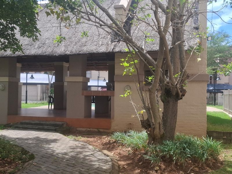 A communal clubhouse area featuring a thatched roof, masonry pillars, and a built-in bar counter, set within a landscaped garden with paved pathways.