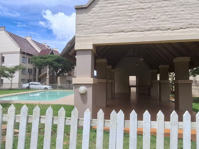 A communal outdoor area featuring a swimming pool and a covered thatched pavilion with brick pillars, set within a landscaped apartment complex.