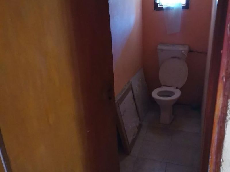 A small, dated bathroom featuring a standard toilet, tiled flooring, and a window. The space requires cosmetic updates and improved lighting.