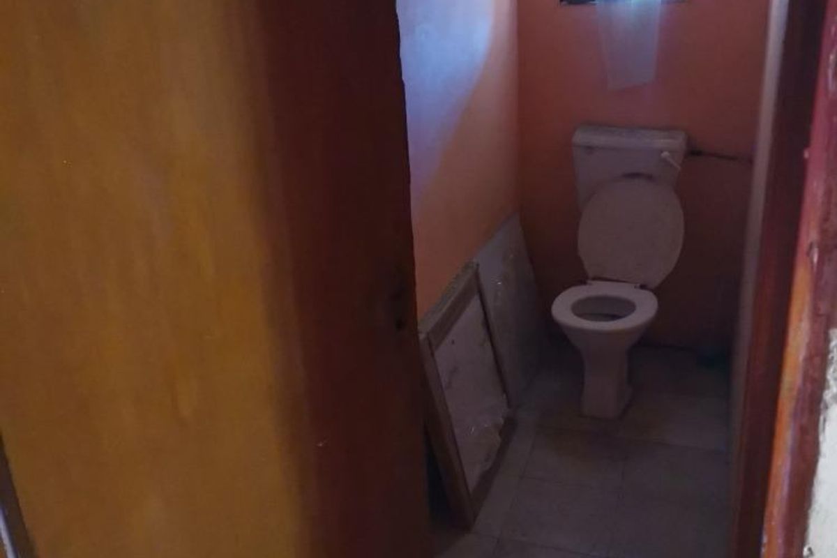 A small, dated bathroom featuring a standard toilet, tiled flooring, and a window. The space requires cosmetic updates and improved lighting.