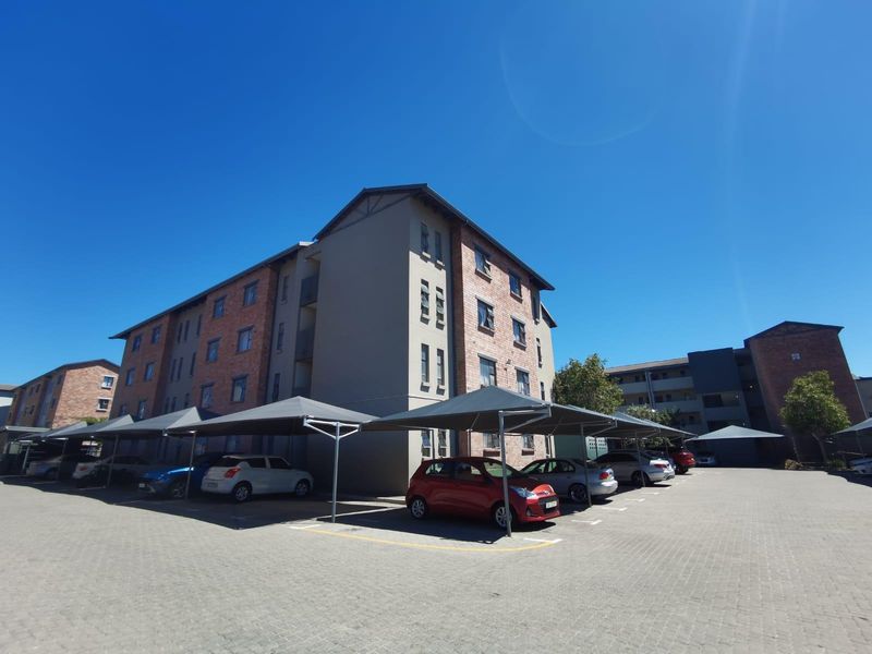 Exterior view of a multi-story apartment complex featuring paved parking bays with shade cloth carports and a brick and plaster facade.