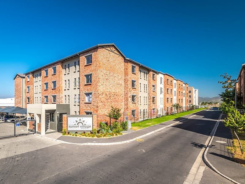 Exterior view of the Sunset Village apartment complex featuring a brick facade, secure gated entrance, and paved driveway access.