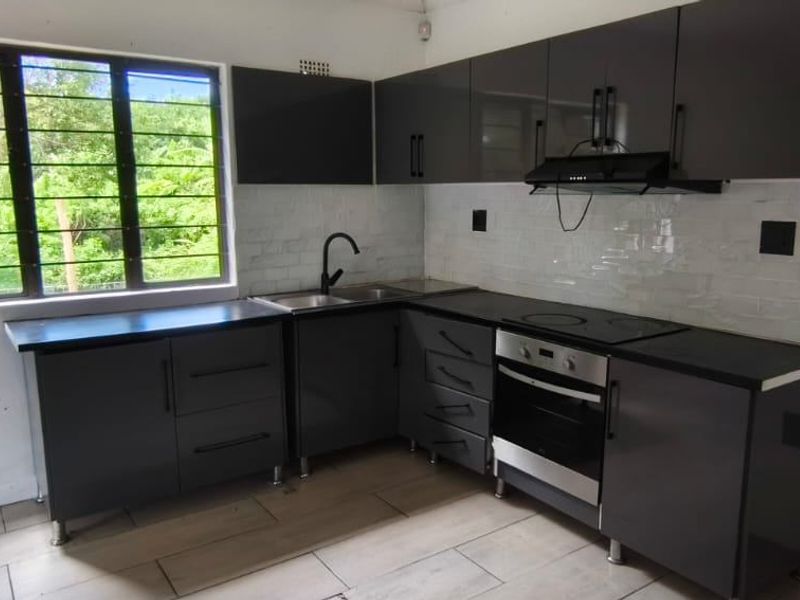 Modern kitchen featuring dark grey cabinetry, integrated oven and hob, tiled backsplash, and a window with security bars overlooking the garden.