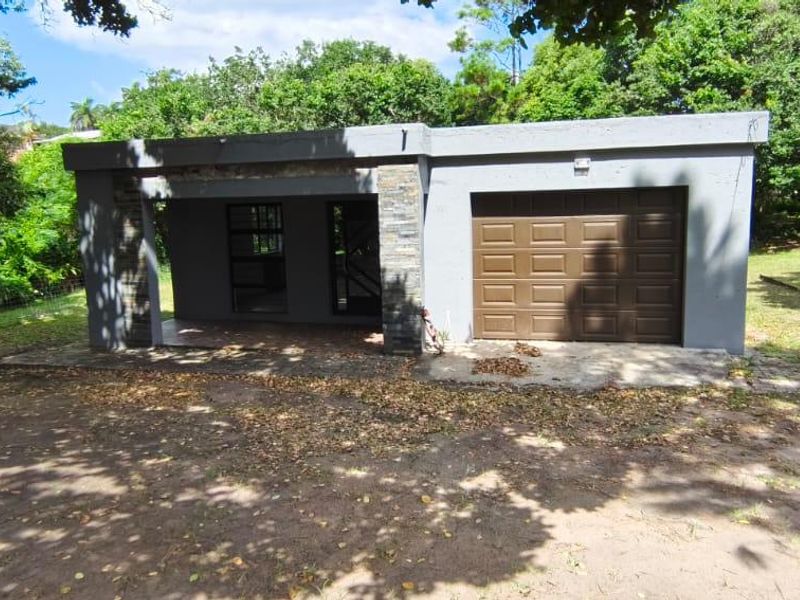 Front elevation of a single-story house featuring a garage, stone-clad pillars, and a covered entrance area set within a wooded garden.