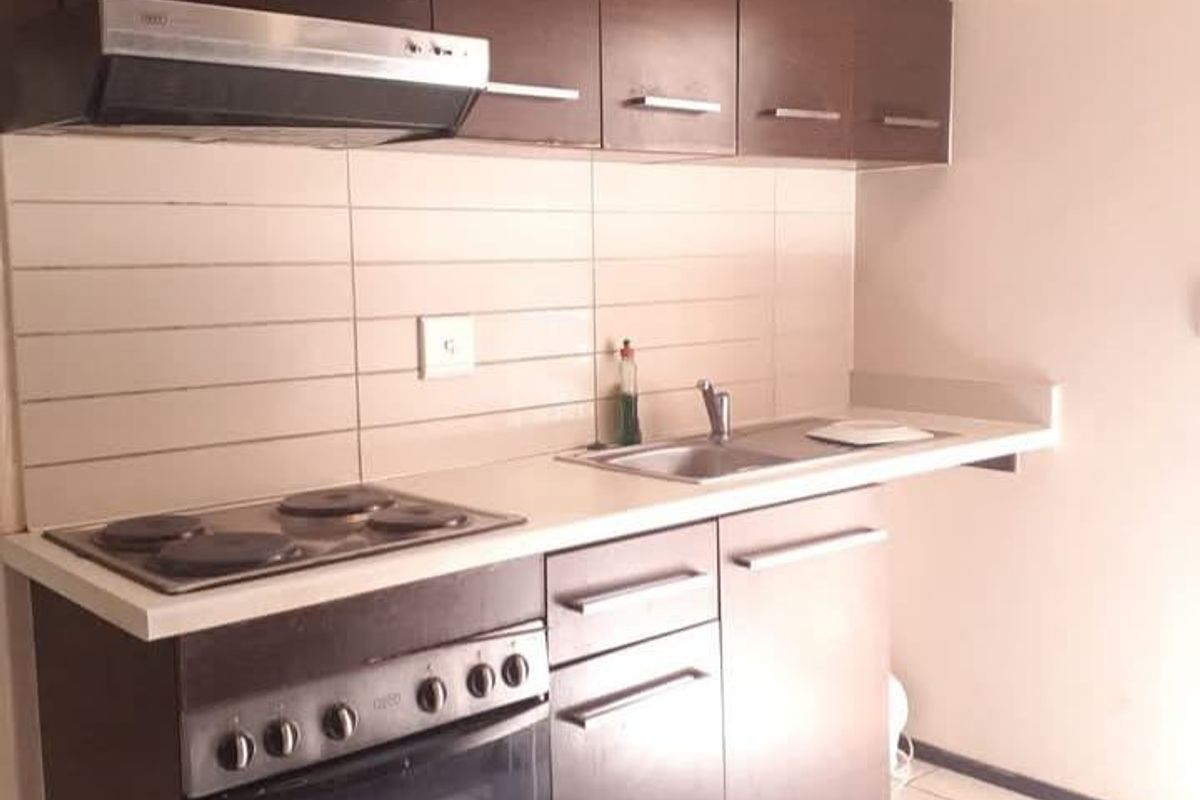 A compact kitchen featuring dark wood cabinetry, built-in oven, electric hob, extractor fan, and tiled splashback with a stainless steel sink.