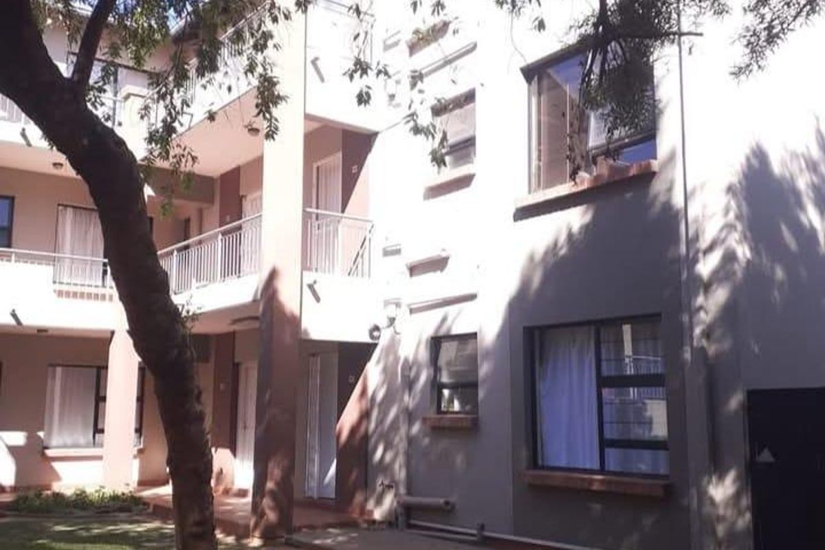 Rear elevation of a multi-story townhouse complex featuring paved walkways, balconies with metal railings, and aluminum-framed windows set within a landscaped courtyard area.