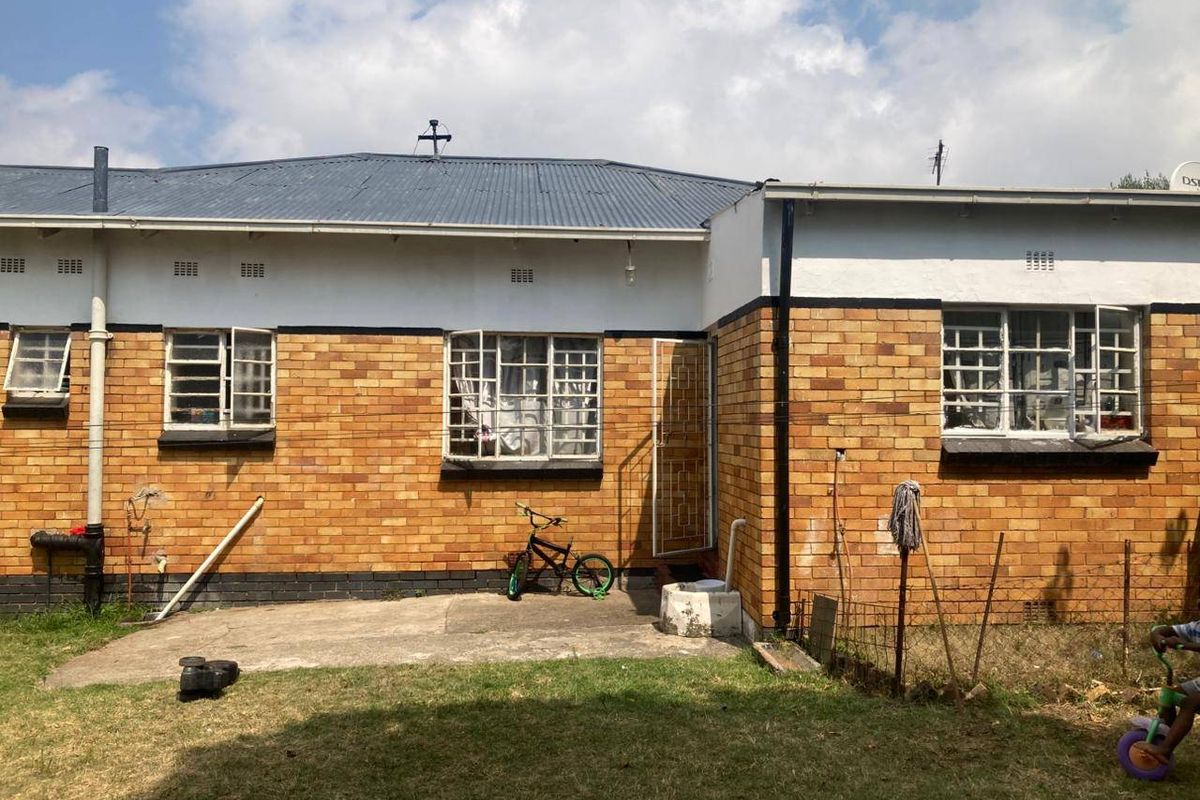 Rear view of a single-story brick house featuring a corrugated metal roof, security-barred windows, and a concrete patio area.