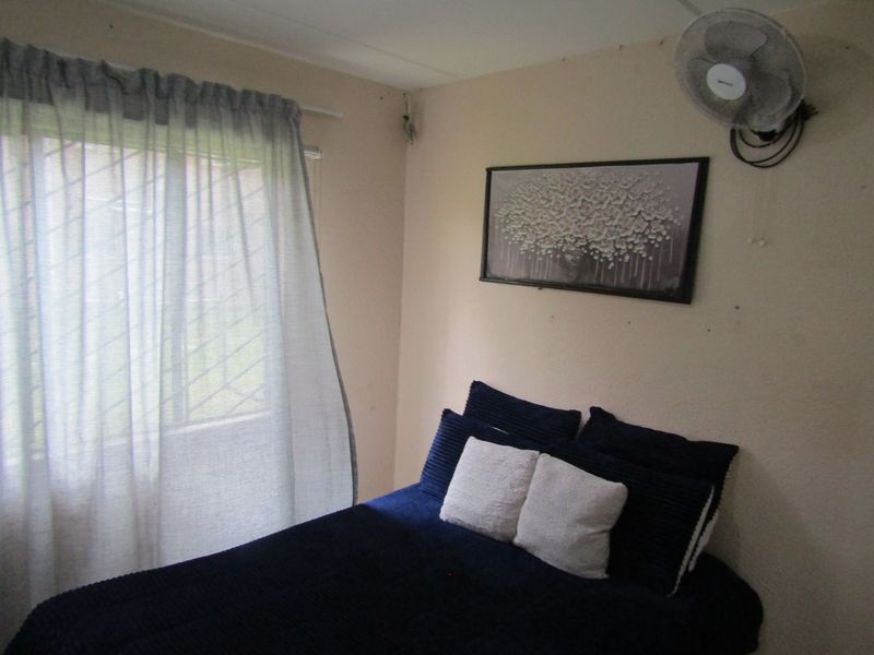 A compact bedroom featuring a wall-mounted fan and a window fitted with security bars.