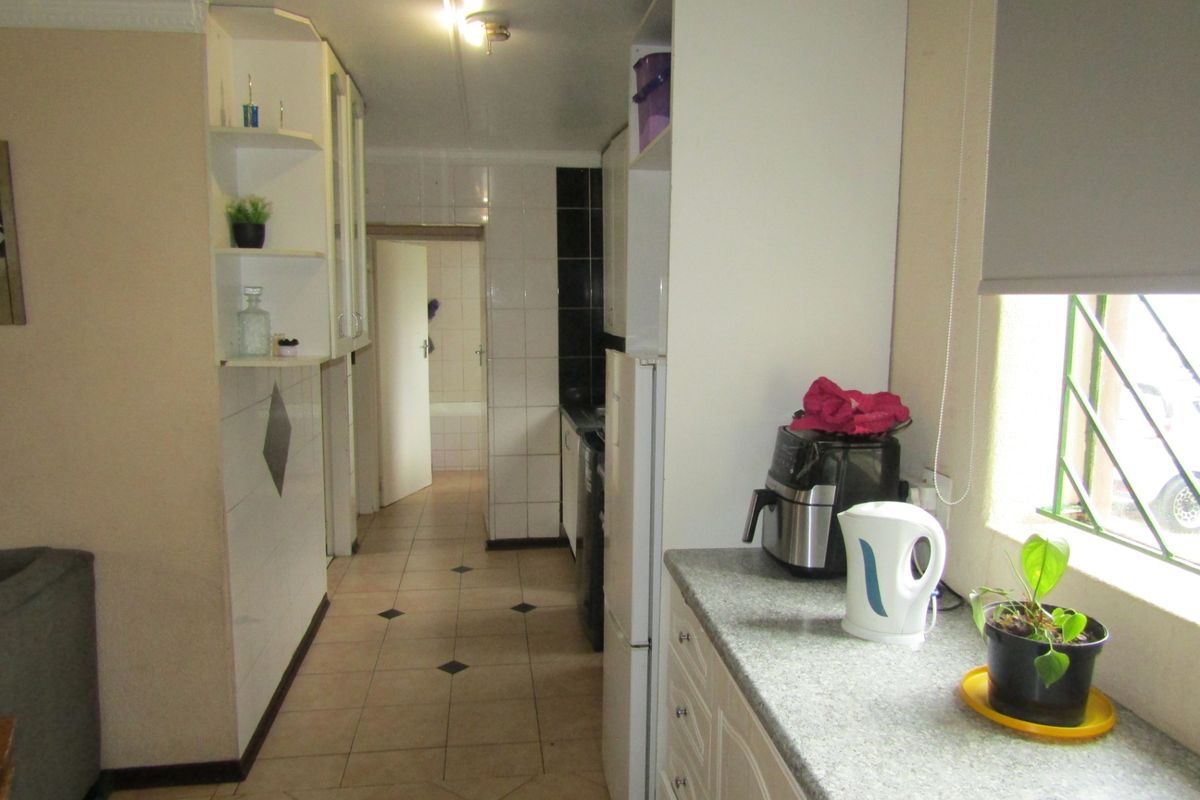 A functional kitchen featuring built-in cabinetry, tiled flooring, and a window with security bars, leading through to an adjacent bathroom area.