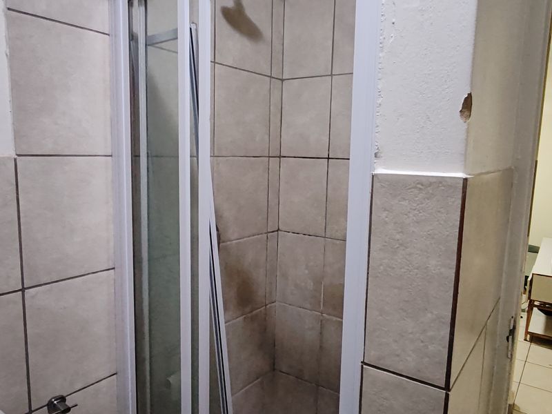 A bathroom featuring a shower enclosure with tiled walls, currently showing signs of wear and damage to the door frame and wall plaster.