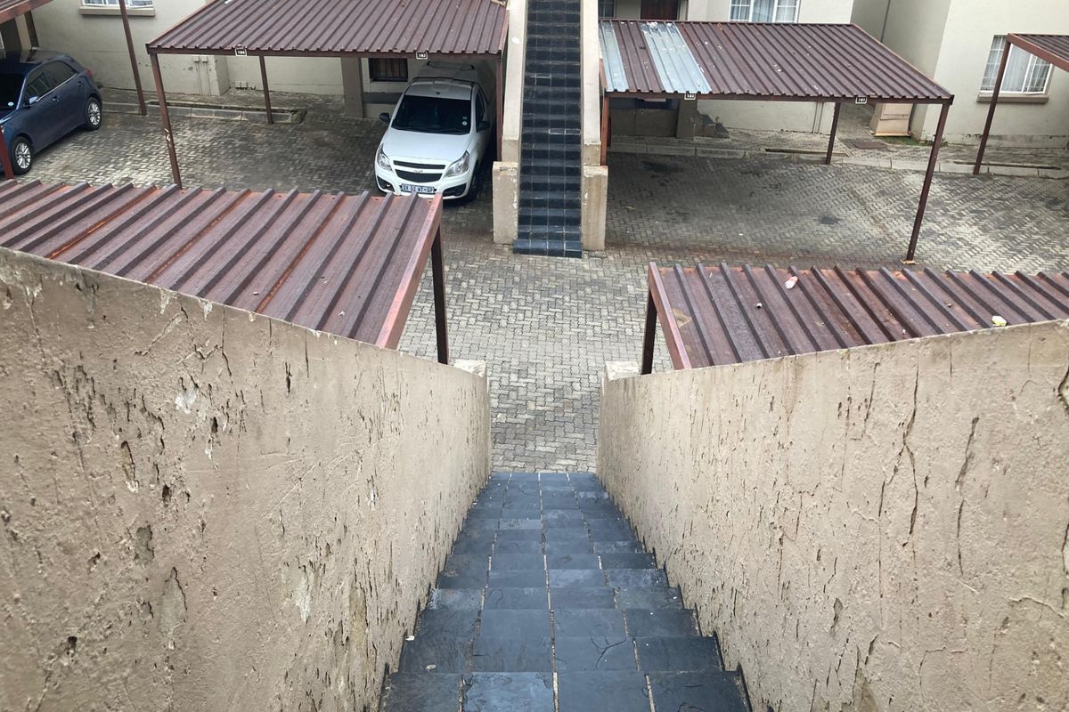 Exterior view of an apartment complex parking area featuring covered carports, paved driveway, and an external staircase with visible wear to the wall finishes.