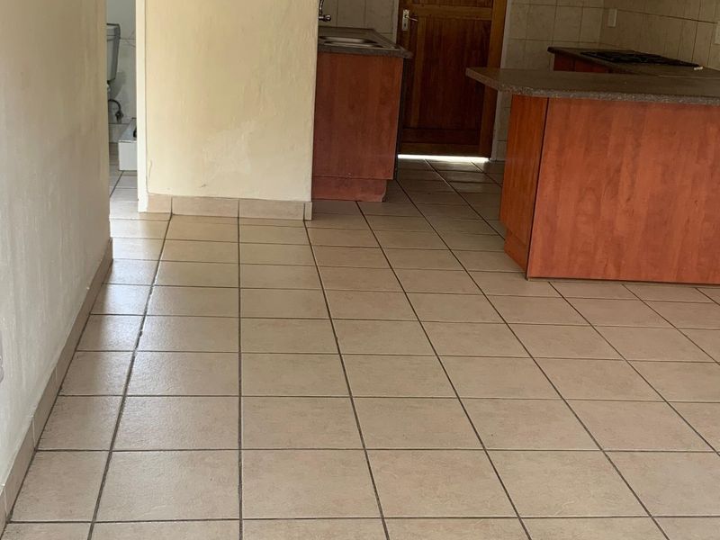 Open plan kitchen area featuring tiled flooring, wooden cabinetry, and a breakfast bar, with an adjacent hallway leading to a bathroom.