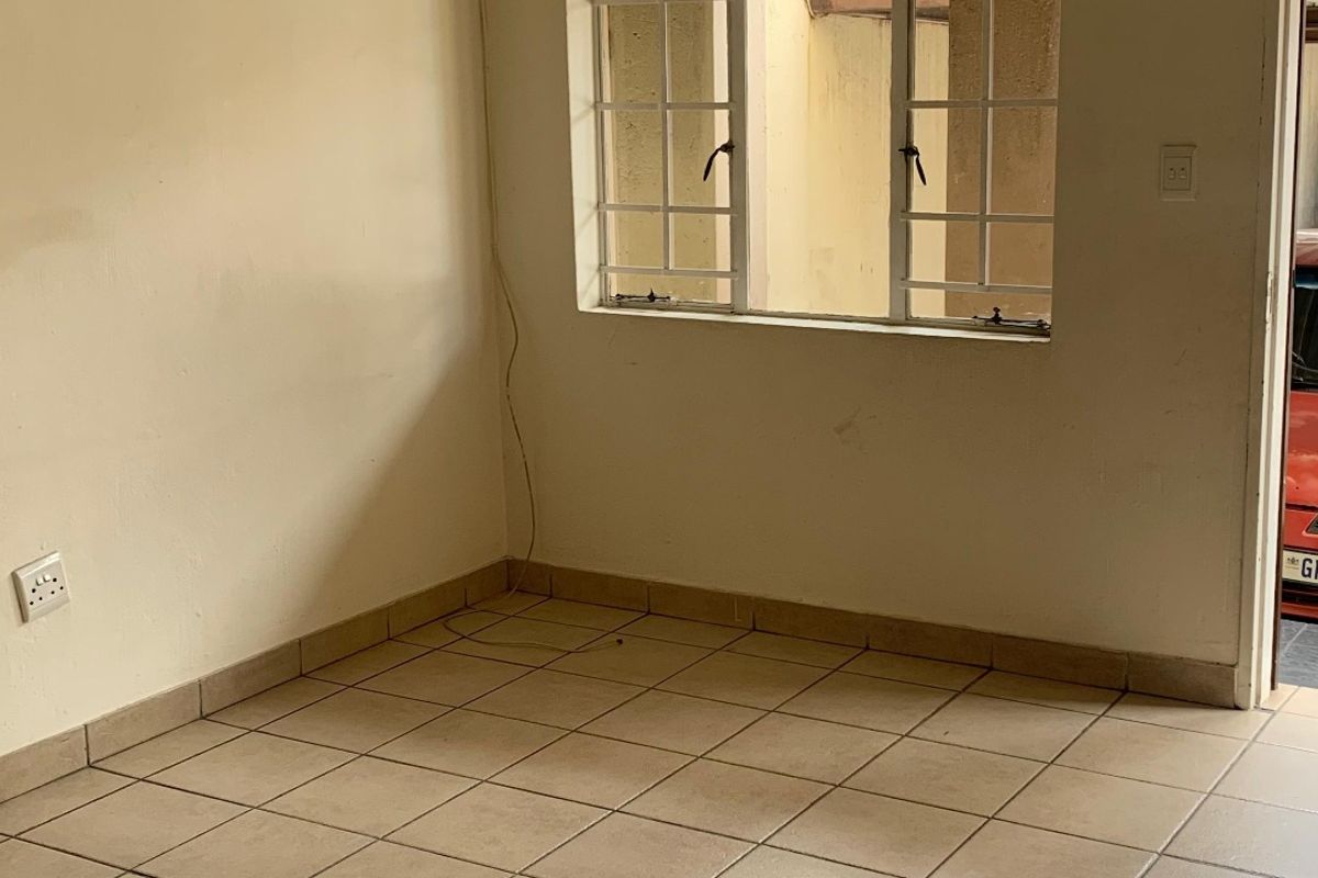 An empty living area featuring neutral ceramic floor tiles, a ceiling-mounted spotlight, and a window with security bars.