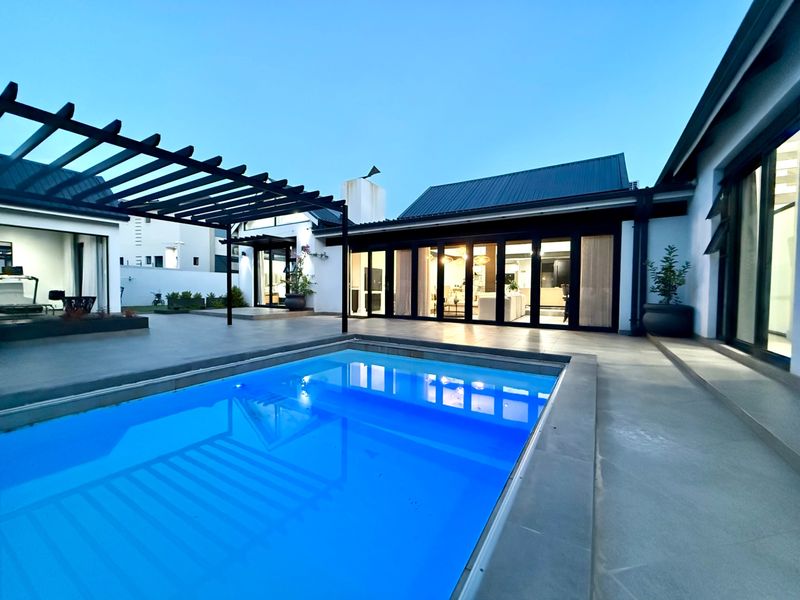 A modern outdoor entertainment area featuring a swimming pool, a timber pergola, and large sliding glass doors leading into the home.