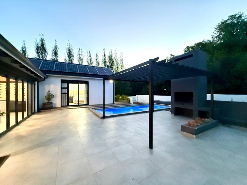 Modern outdoor entertainment area featuring a swimming pool, a covered pergola with a built-in braai, and large sliding doors leading to the interior.
