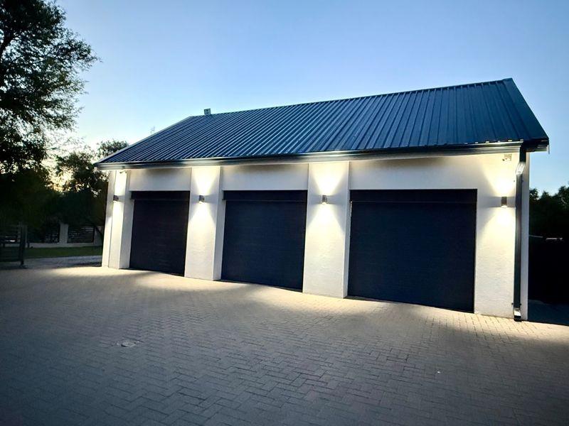 A well-maintained triple garage featuring modern dark roller doors, a metal roof, and integrated exterior wall lighting, set against a paved driveway.