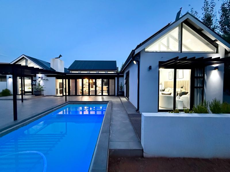 A beautifully illuminated modern home exterior featuring a swimming pool, paved patio area, and large glass sliding doors connecting indoor and outdoor living spaces.