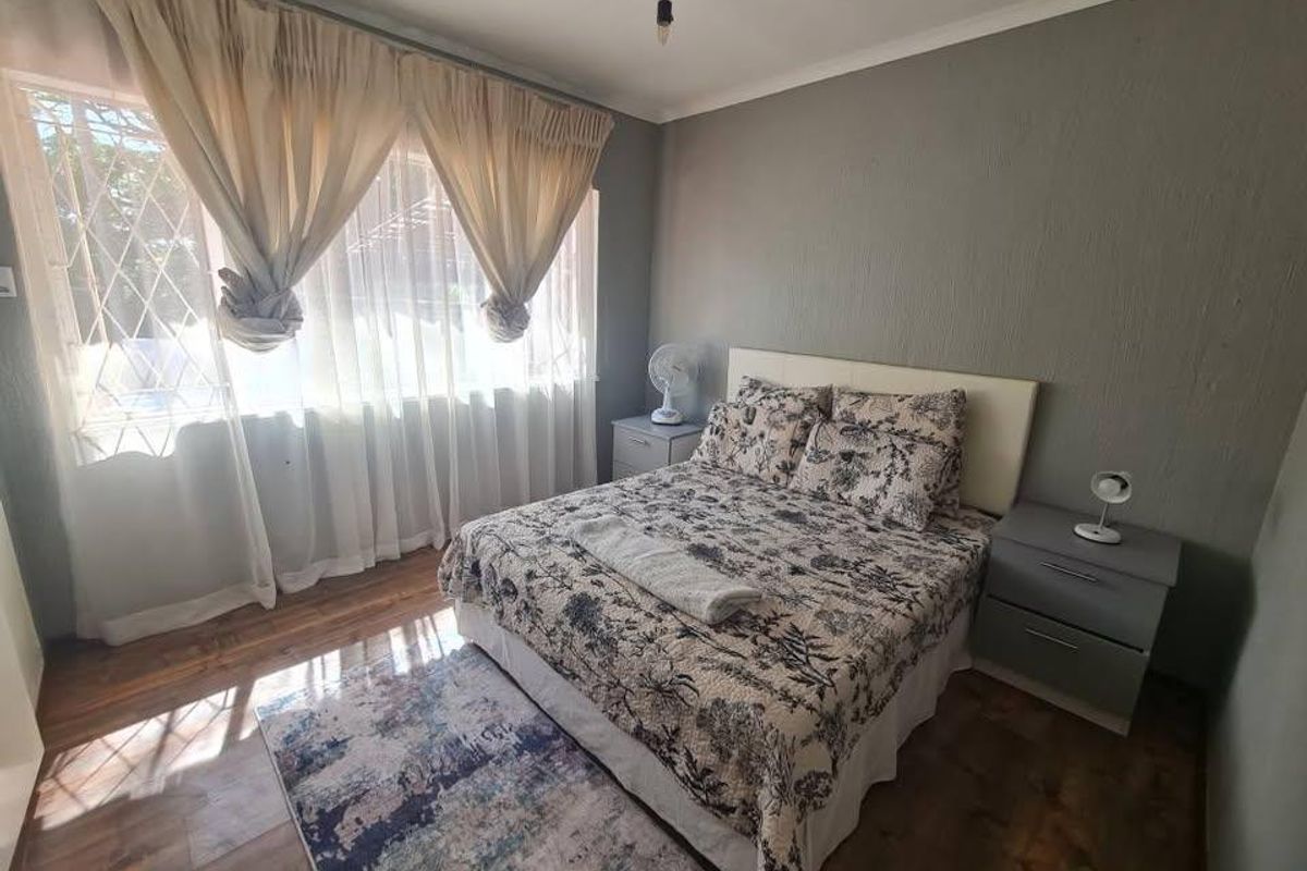 A well-lit bedroom featuring laminate flooring, neutral grey walls, and large windows fitted with security bars.
