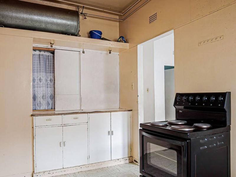 A dated kitchen featuring original built-in cabinetry, an electric stove, and an exposed hot water cylinder, requiring cosmetic updates and modernization.