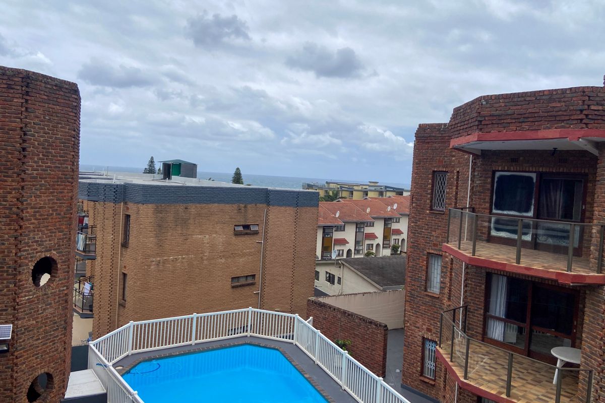 Elevated view of a communal swimming pool area surrounded by brick apartment buildings with balconies and distant ocean views.