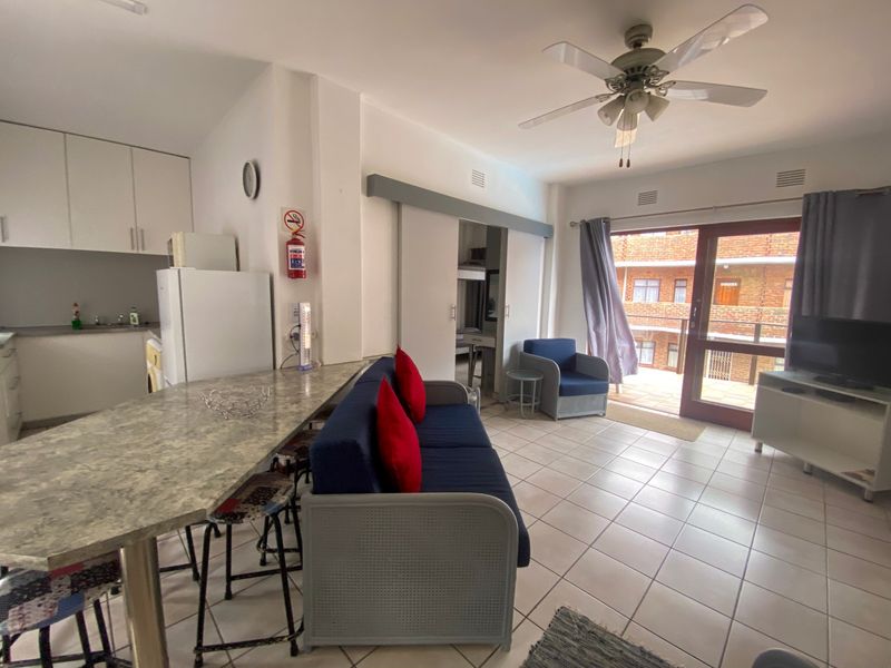 Open-plan living area featuring a kitchen with built-in cabinetry, tiled flooring, a ceiling fan, and direct access to a balcony through a sliding door.